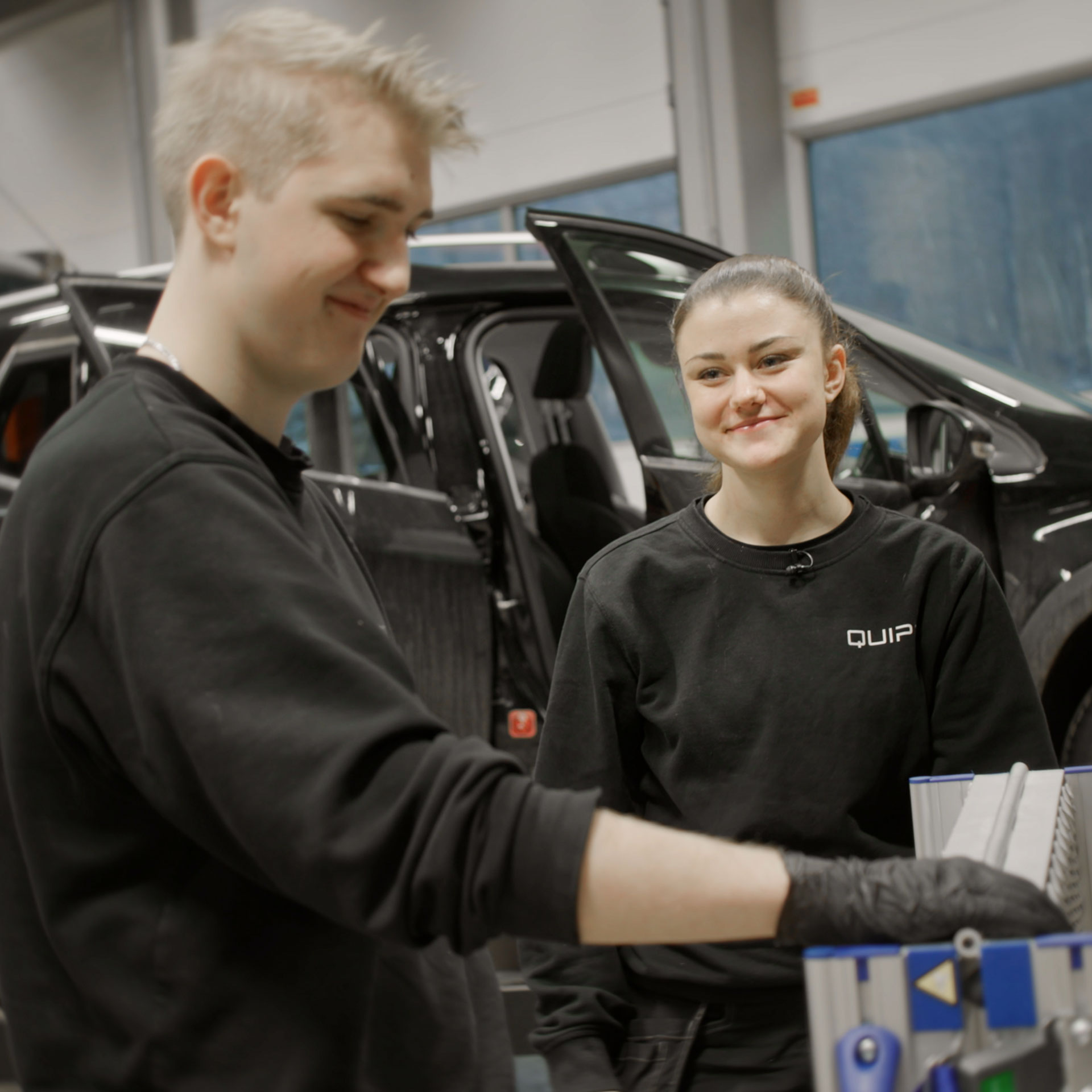 To unge bilteknikere arbeider sammen i et verksted, med en bil i bakgrunnen – et innblikk i arbeidshverdagen for lærlinger hos QUIPD.